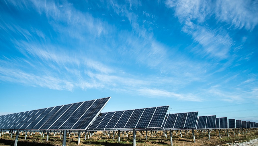 Rows of tilted solar panels on a utility-scale solar farm, mounted on green-yellow grass, with a light blue sky and white clouds above.
