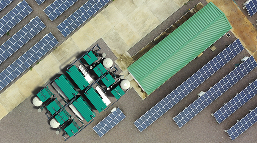 A 3D-rendered top view shows rows of solar panels, a building with green roof and a storage area with green containers, white tanks and steel pipes connecting them.