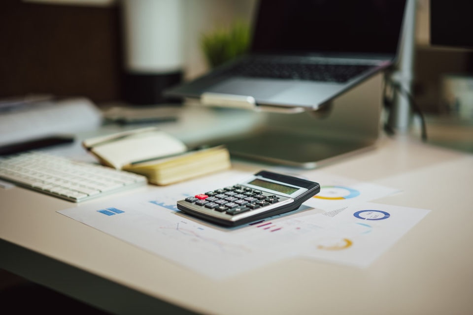 A calculator and sheets of paper on a desk, with a laptop in the background